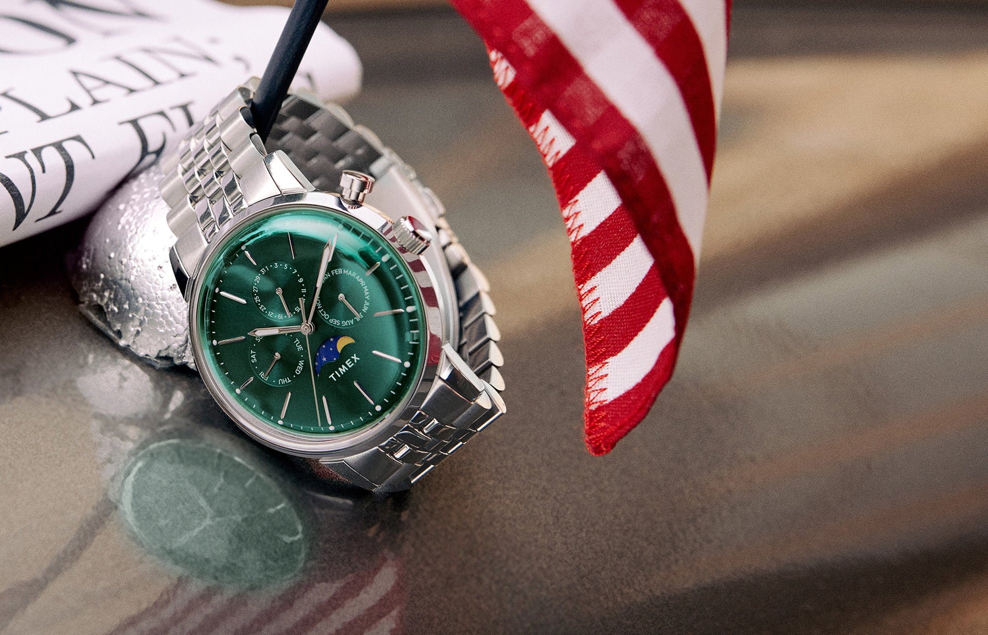 Timex Marlin Moon Phase Multifunction with deep green dial and stainless-steel bracelet placed beside a small American flag on a reflective surface.