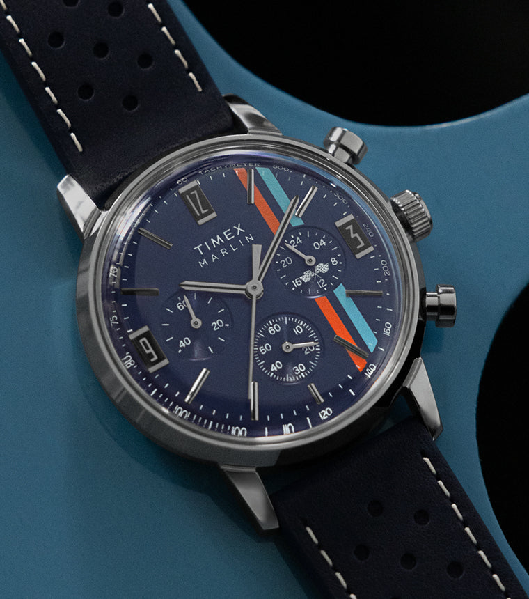 Timex Marlin Chronograph Tachymeter with navy dial, multi-color racing stripes, and perforated blue leather strap resting on a blue metal surface.