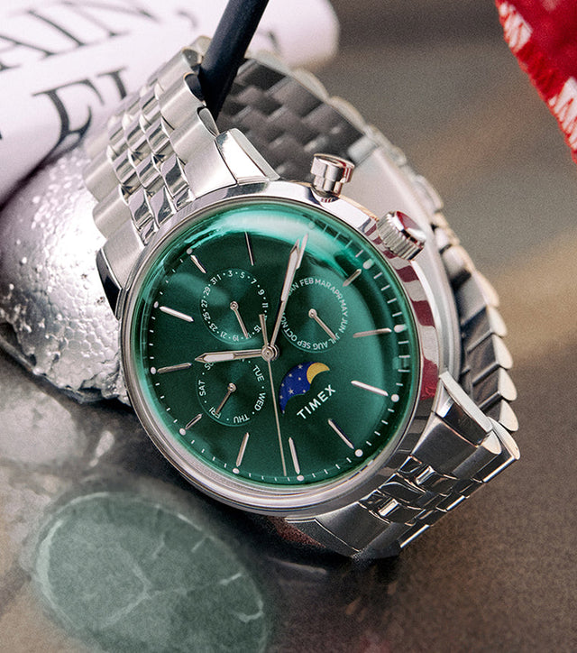 Timex Marlin Moon Phase Multifunction with deep green dial and stainless-steel bracelet placed beside a small American flag on a reflective surface.