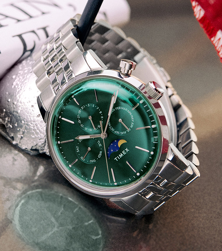 Timex Marlin Moon Phase Multifunction with deep green dial and stainless-steel bracelet placed beside a small American flag on a reflective surface.