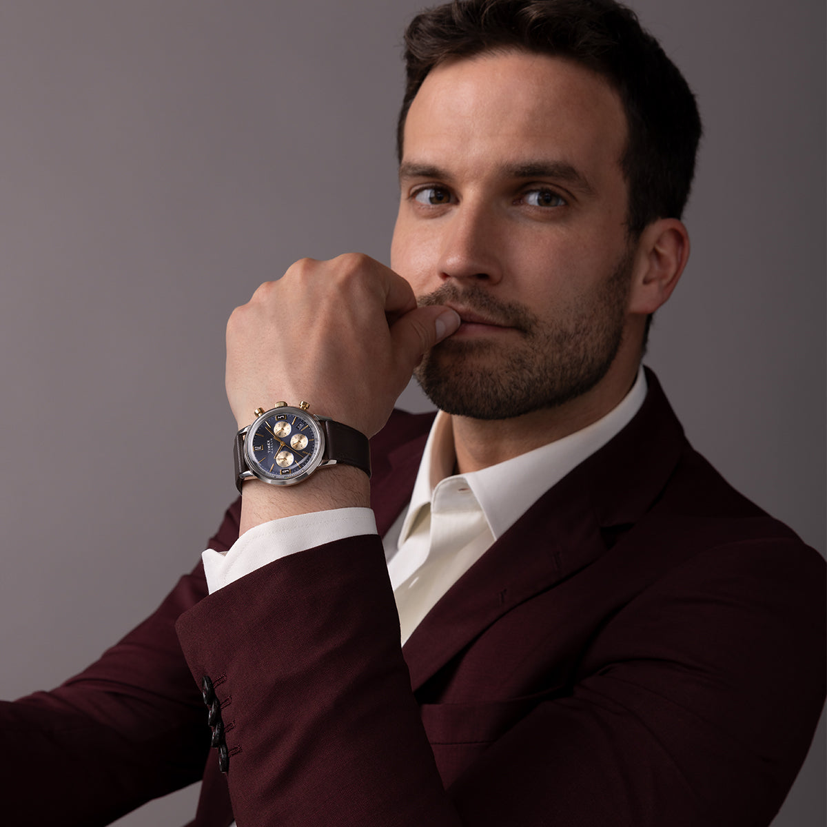 Model wearing Timex Marlin Chronograph Tachymeter with blue dial and brown leather strap, dressed in a burgundy suit.