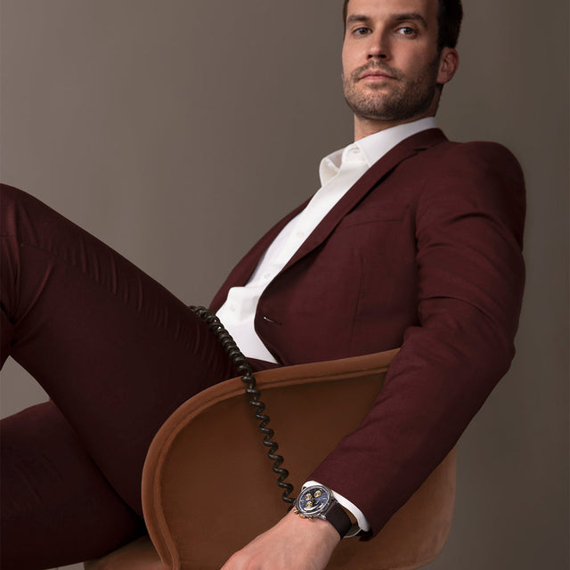 Model seated in a chair wearing Timex Marlin Chronograph Tachymeter with navy dial and brown strap, paired with a burgundy suit.