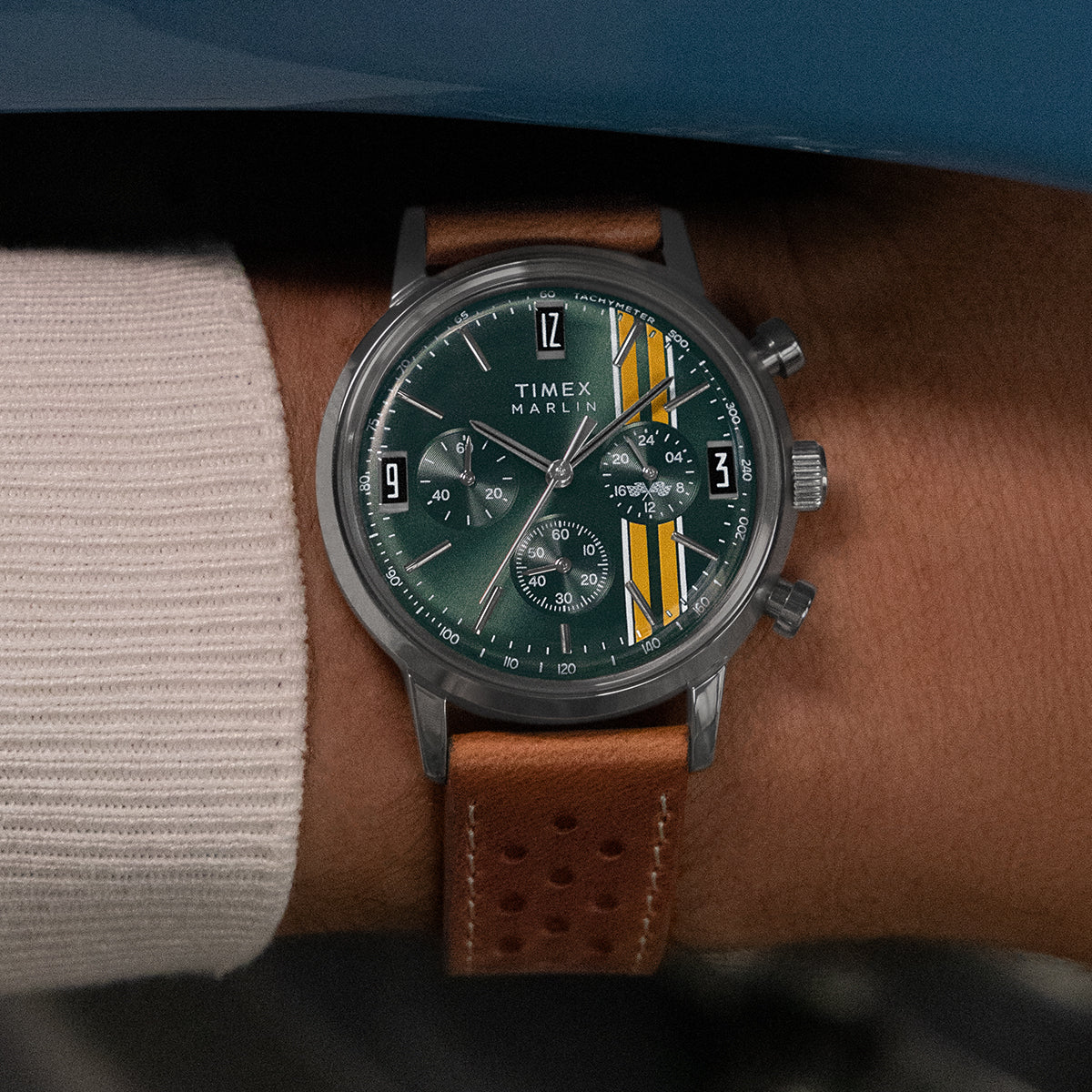 Close-up of model wearing Timex Marlin Chronograph Tachymeter with green dial, yellow accents, and brown strap beside a vintage blue race car.
