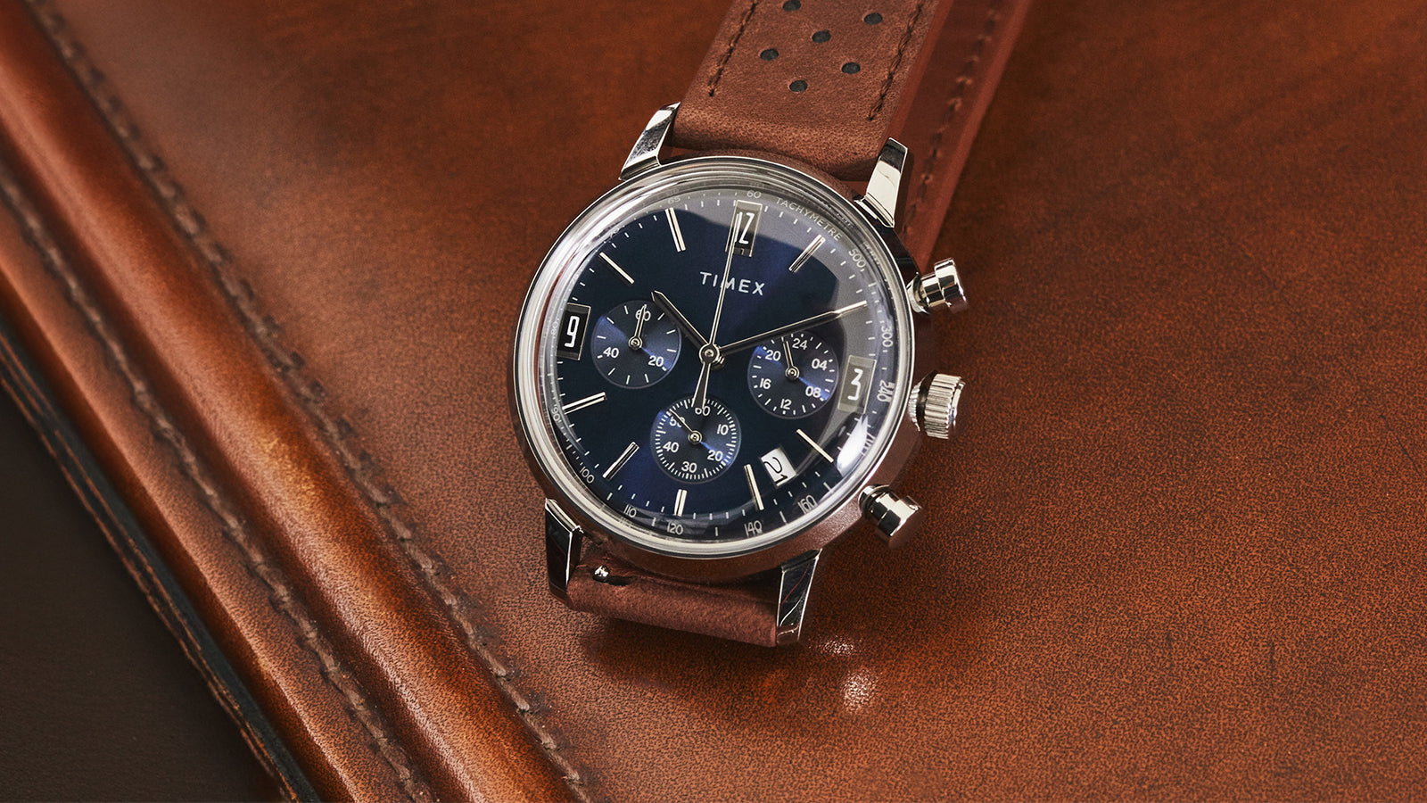 Timex Marlin Chronograph Tachymeter watch with blue dial and brown perforated leather strap resting on brown leather surface.