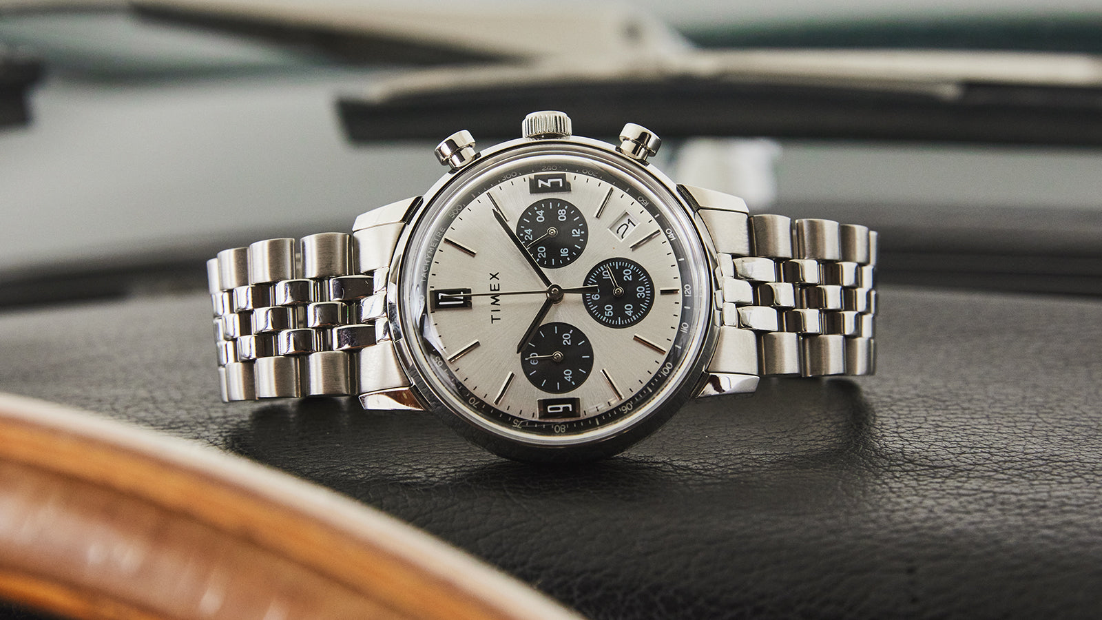 Timex Marlin Chronograph Tachymeter with silver dial and stainless-steel bracelet on black leather car interior background.