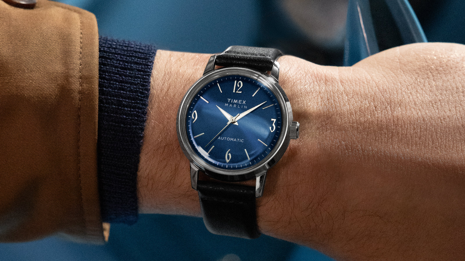 Timex Marlin Automatic watch with blue dial and silver-tone markers worn on wrist, paired with a brown jacket and navy knit cuff.