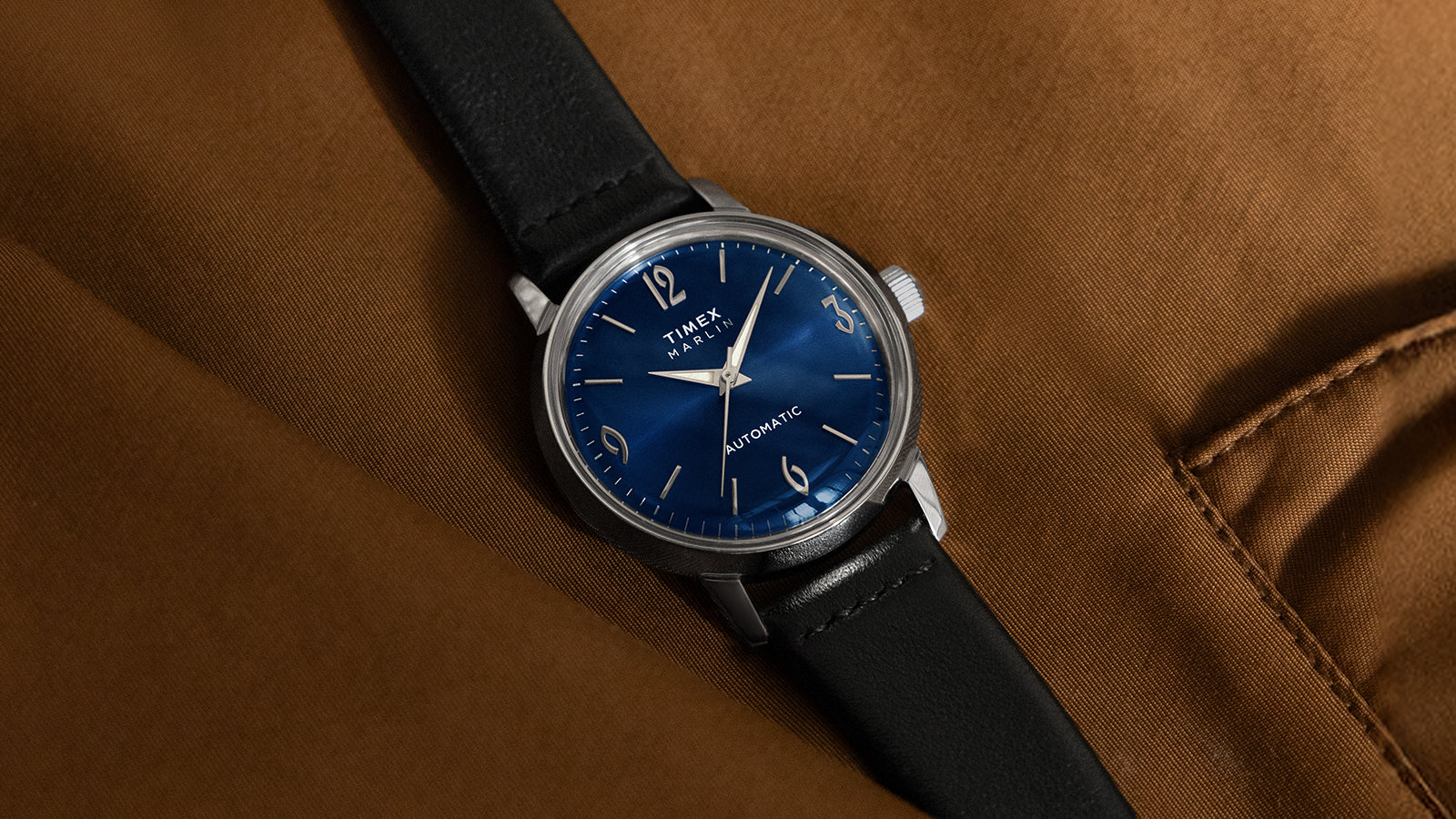 Timex Marlin Automatic watch with deep blue sunray dial and black leather strap resting on a brown fabric surface.