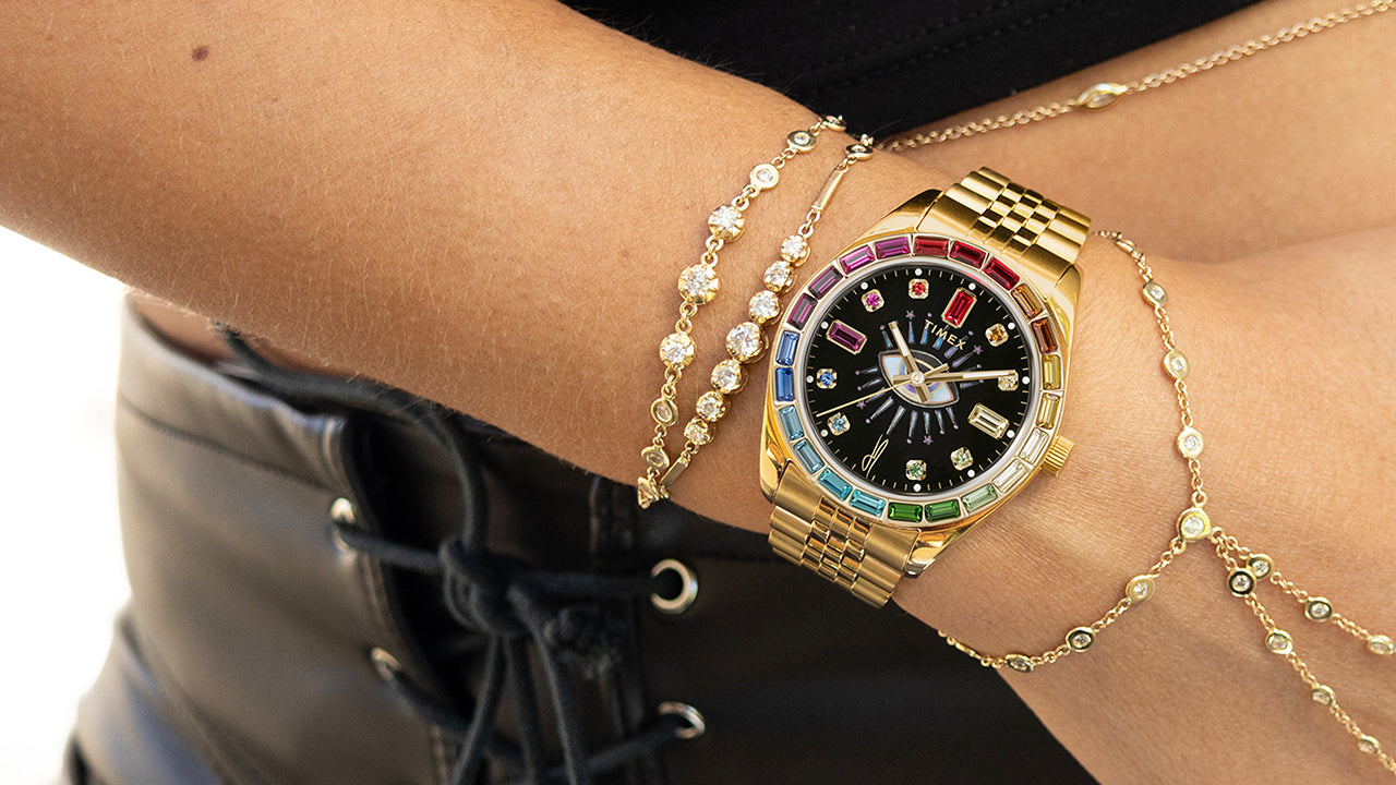 Timex x Jacquie Aiche “Cosmic Nights” watch with a gold-tone case and bracelet featuring multicolored gemstone accents. The black dial displays crystal hour markers and a radiant “all-seeing eye” motif at the center, blending celestial and luxury design elements.