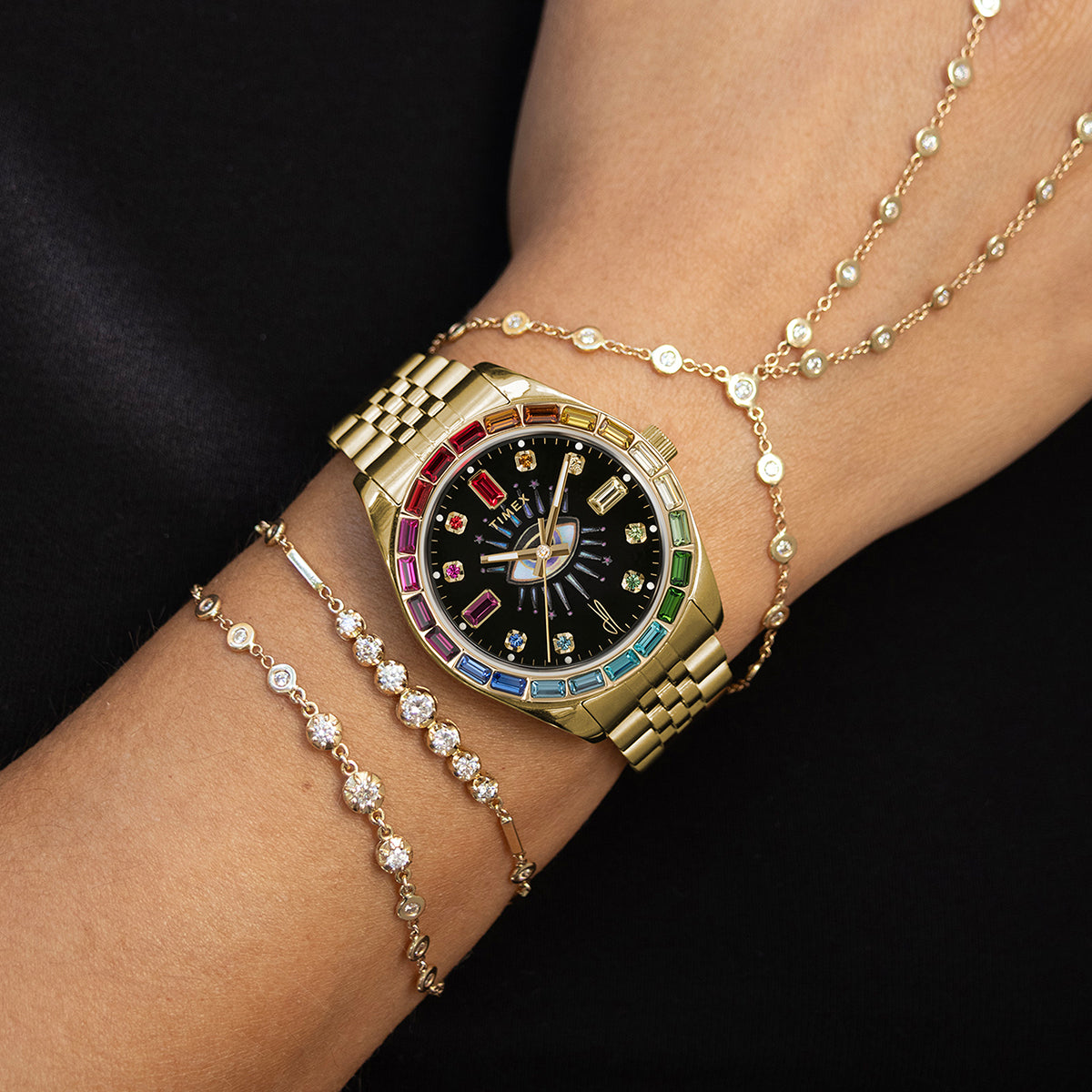 Lifestyle shot of the Timex x Jacquie Aiche “Cosmic Nights” watch worn on the wrist with layered gold and diamond bracelets. The gold case and colorful bezel sparkle under natural light, highlighting its bohemian-luxury style.