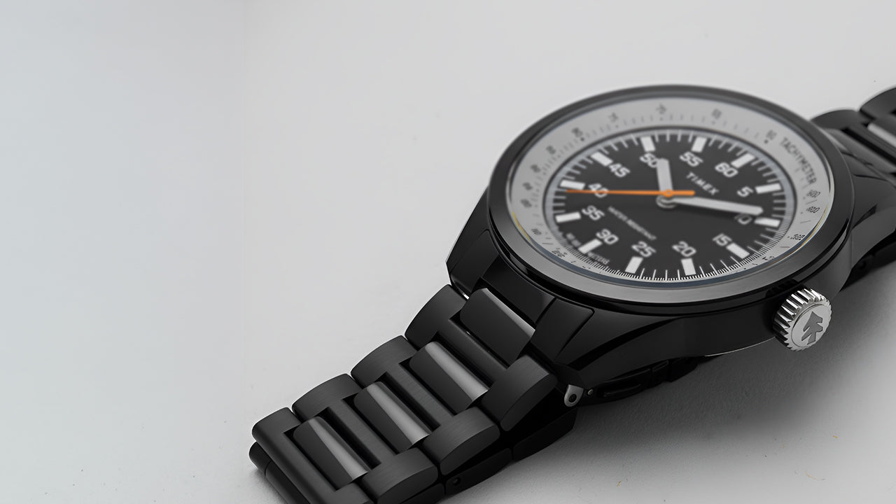 A Huckberry x Timex 1979 Black Max watch on a clean, light gray background. The black metal bracelet and case contrast with the white outer dial ring and black center dial, highlighted by an orange seconds hand.