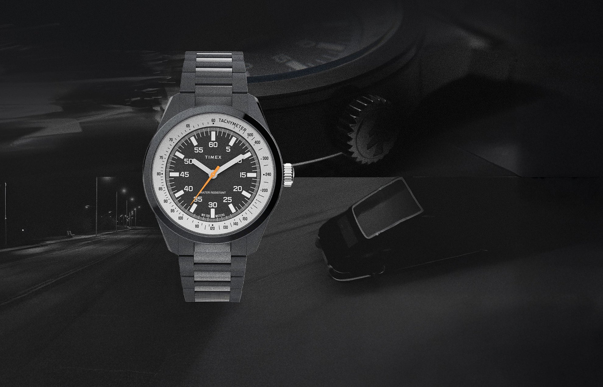 The Huckberry x Timex 1979 Black Max watch displayed against a dark, moody background featuring a night road and a faint silhouette of a car. The watch has a black case and bracelet, a black dial with white numerals, an orange seconds hand, and a tachymeter scale.