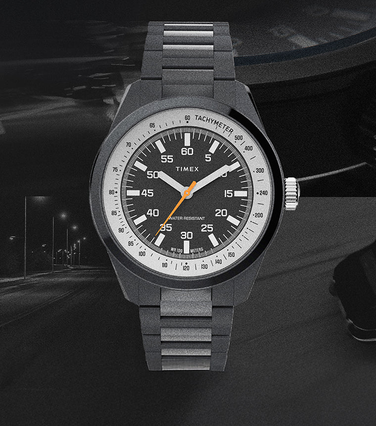 The Huckberry x Timex 1979 Black Max watch displayed against a dark, moody background featuring a night road and a faint silhouette of a car. The watch has a black case and bracelet, a black dial with white numerals, an orange seconds hand, and a tachymeter scale.