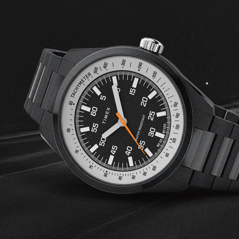 Huckberry x Timex field-style watch with a black dial, white tachymeter ring, orange seconds hand, and black case and bracelet, shown in a close-up against a dark textured background.