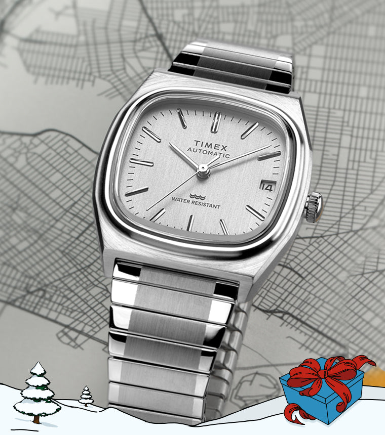 Timex Automatic 1983 E Line Reissue stainless steel watch with a silver dial and bracelet, displayed against a grayscale city map background. The bottom foreground features an illustrated snowy landscape with pine trees and a blue gift box wrapped with a red ribbon, evoking a winter holiday theme.
