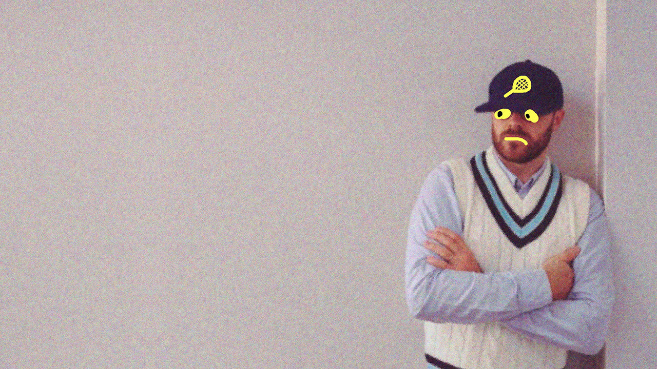 A portrait of a man standing with arms crossed against a neutral wall, wearing a white cricket-style sweater and cap with playful doodles drawn on his face and hat.