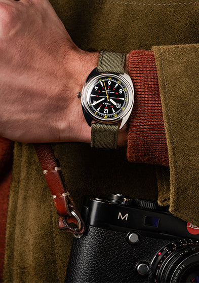 Lifestyle image of the Timex x Bespoke Post watch worn on a wrist, paired with a brown suede jacket and camera strap. The shot captures rugged sophistication and adventure-ready elegance.