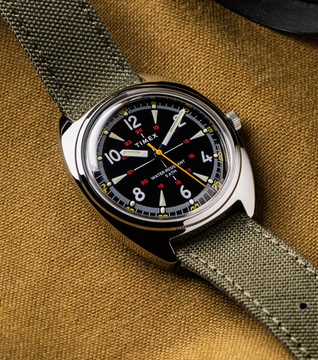 Timex x Bespoke Post watch with a black dial, luminous markers, and yellow accents, paired with an olive green fabric strap. The watch rests on a textured mustard fabric background, evoking rugged, adventure-ready style.