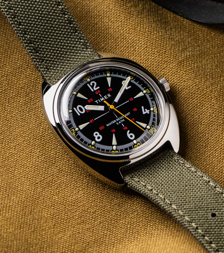 Timex x Bespoke Post watch with a black dial, luminous markers, and yellow accents, paired with an olive green fabric strap. The watch rests on a textured mustard fabric background, evoking rugged, adventure-ready style.