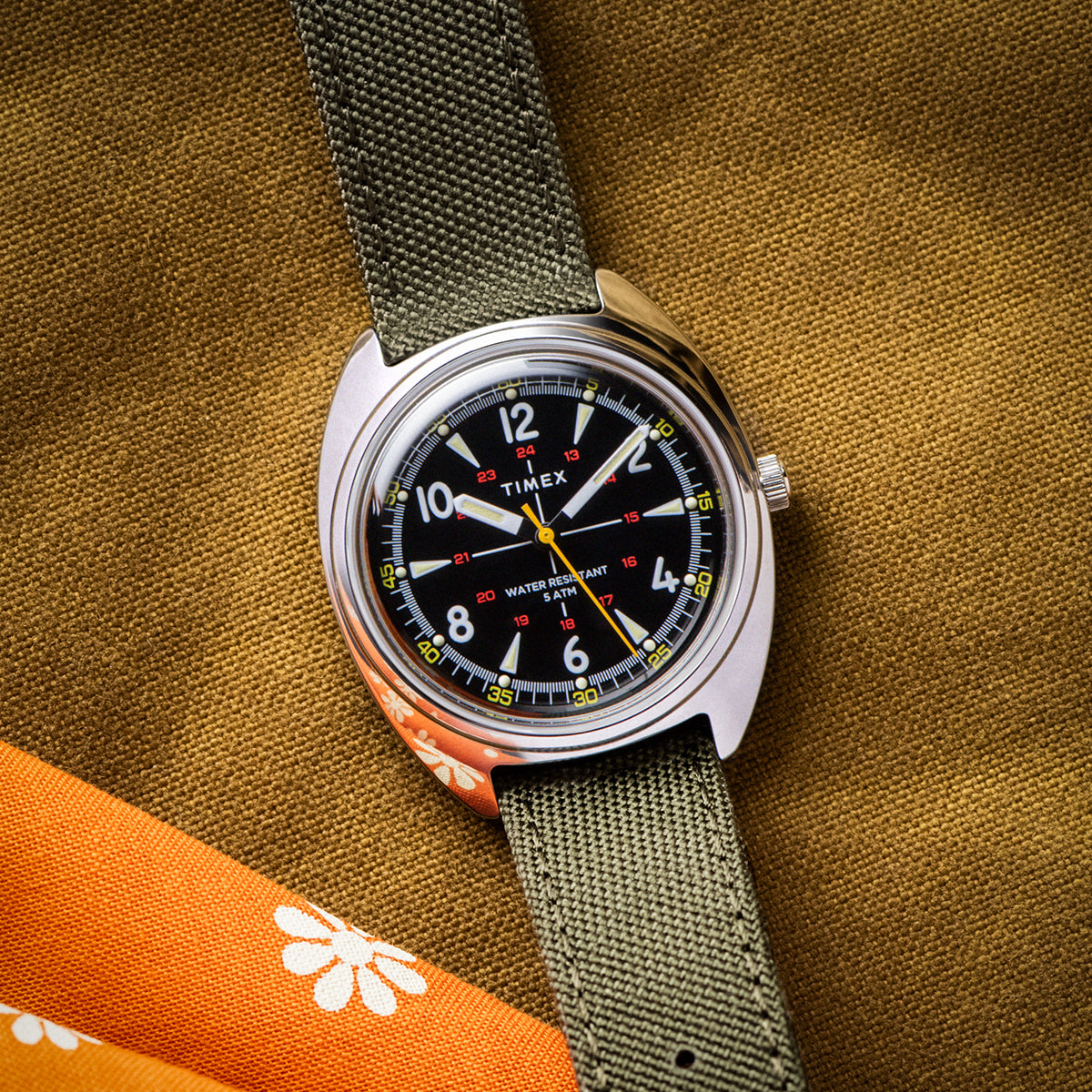 Close-up view of the Timex x Bespoke Post watch with a black dial featuring bold white numerals and red 24-hour markers. The olive strap and brushed steel case highlight its utilitarian and timeless aesthetic.