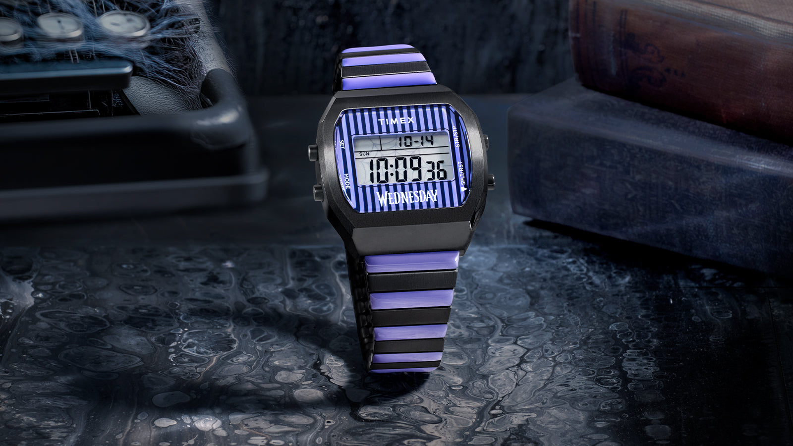 Close-up of the Timex x Wednesday T80 digital watch with a black rectangular case, purple and black striped bracelet, and digital display reading “Wednesday.” The eerie lighting highlights its gothic-inspired design and classic Timex digital layout.