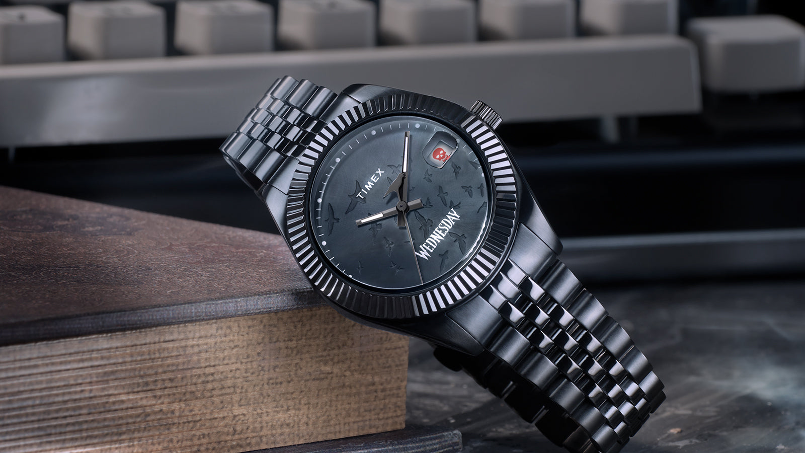 Close-up of the Timex x Wednesday Timex Legacy watch with a brushed black stainless-steel bracelet and fluted bezel. The dial shows embossed flying ravens and gothic typography, evoking the eerie elegance of Wednesday Addams.