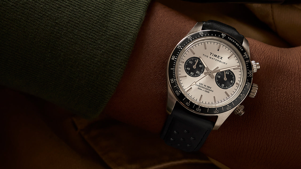 Timex Waterbury Heritage Chronograph on wrist, showing silver-tone dial, black sub-dials, and black perforated leather strap paired with a green sweater.
