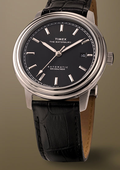 Timex Waterbury Metropolitan Automatic watch featuring a black dial, polished stainless steel case, and black leather strap on a neutral beige background.