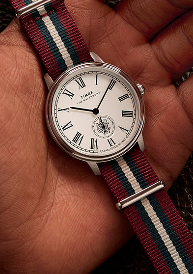 Timex Waterbury Metropolitan Sub-Second held in hand above a brown leather bag, showcasing its classic design and striped fabric strap.