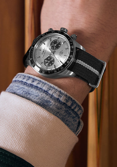 Person wearing the Timex Waterbury Heritage Chronograph with silver and black dial and striped fabric strap while holding a pool cue near a wooden rack.
