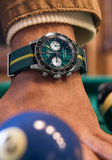 Close-up of the Timex Waterbury Heritage Chronograph with green dial and striped fabric strap worn with a tan jacket sleeve.