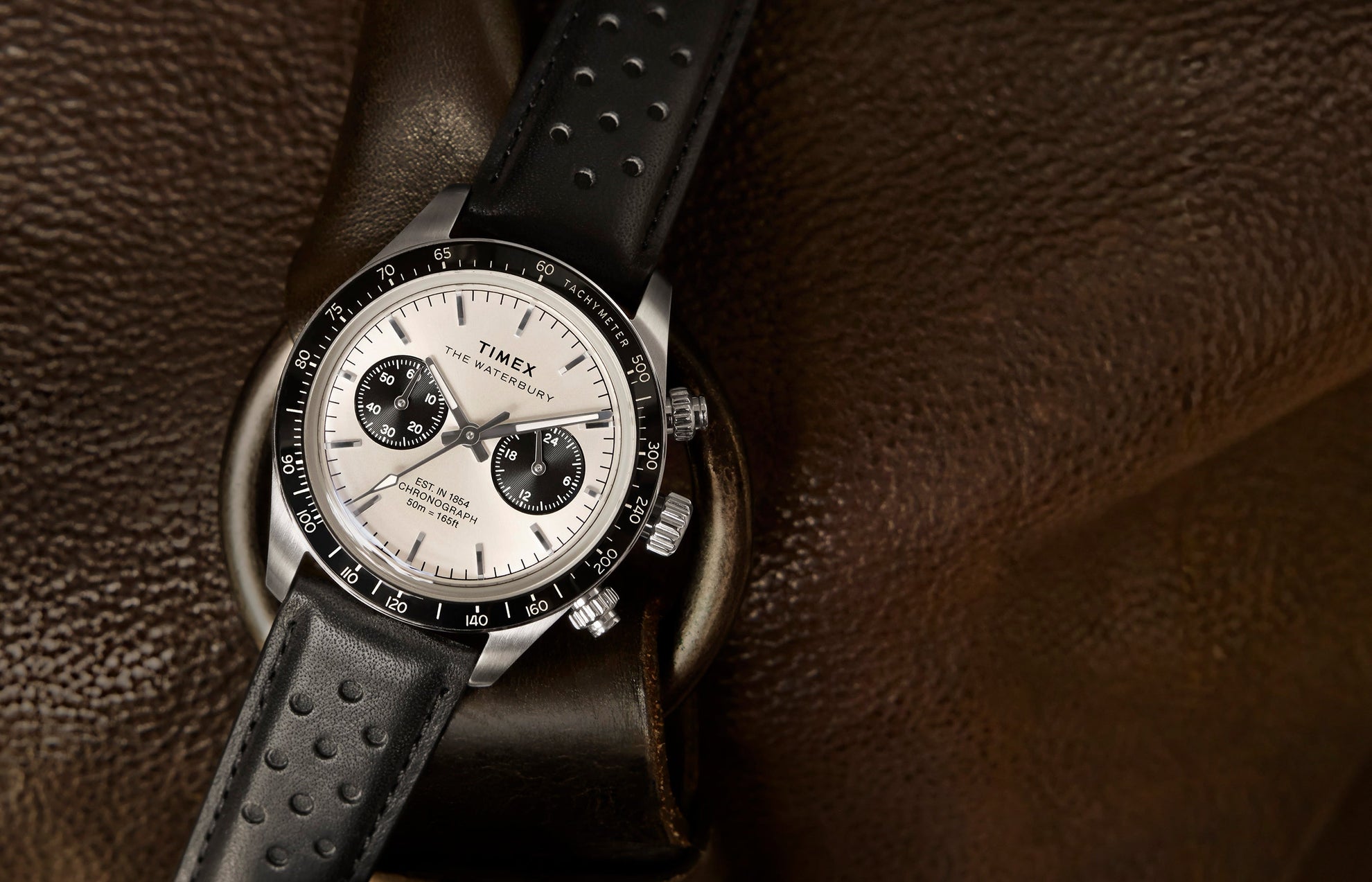 Timex Waterbury Heritage Chronograph with silver dial and black sub-dials resting on brown leather, featuring a black perforated leather strap and tachymeter bezel.