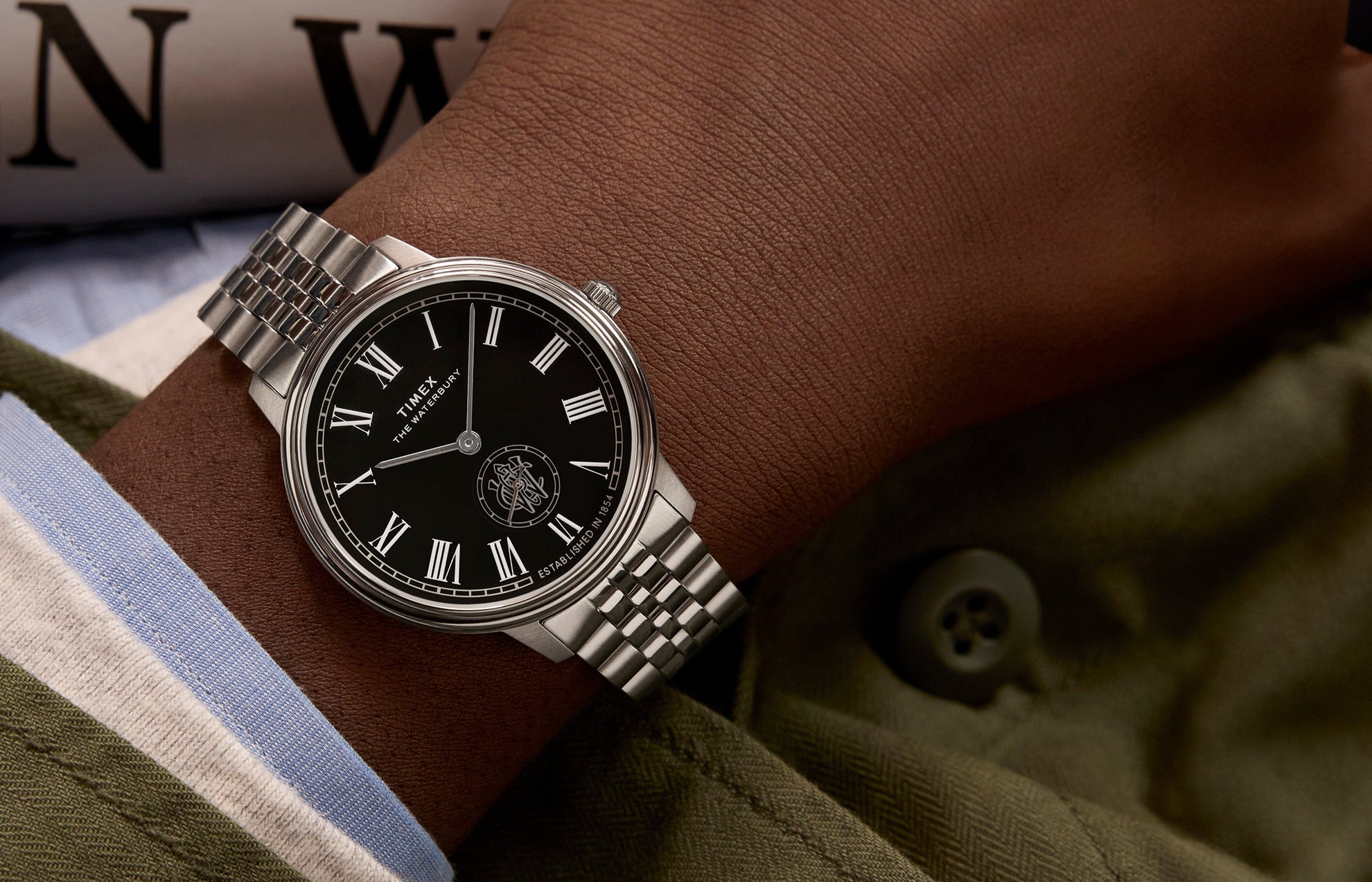Timex Waterbury Metropolitan Sub-Second watch with black dial, Roman numerals, and stainless steel bracelet worn on wrist with a green jacket.