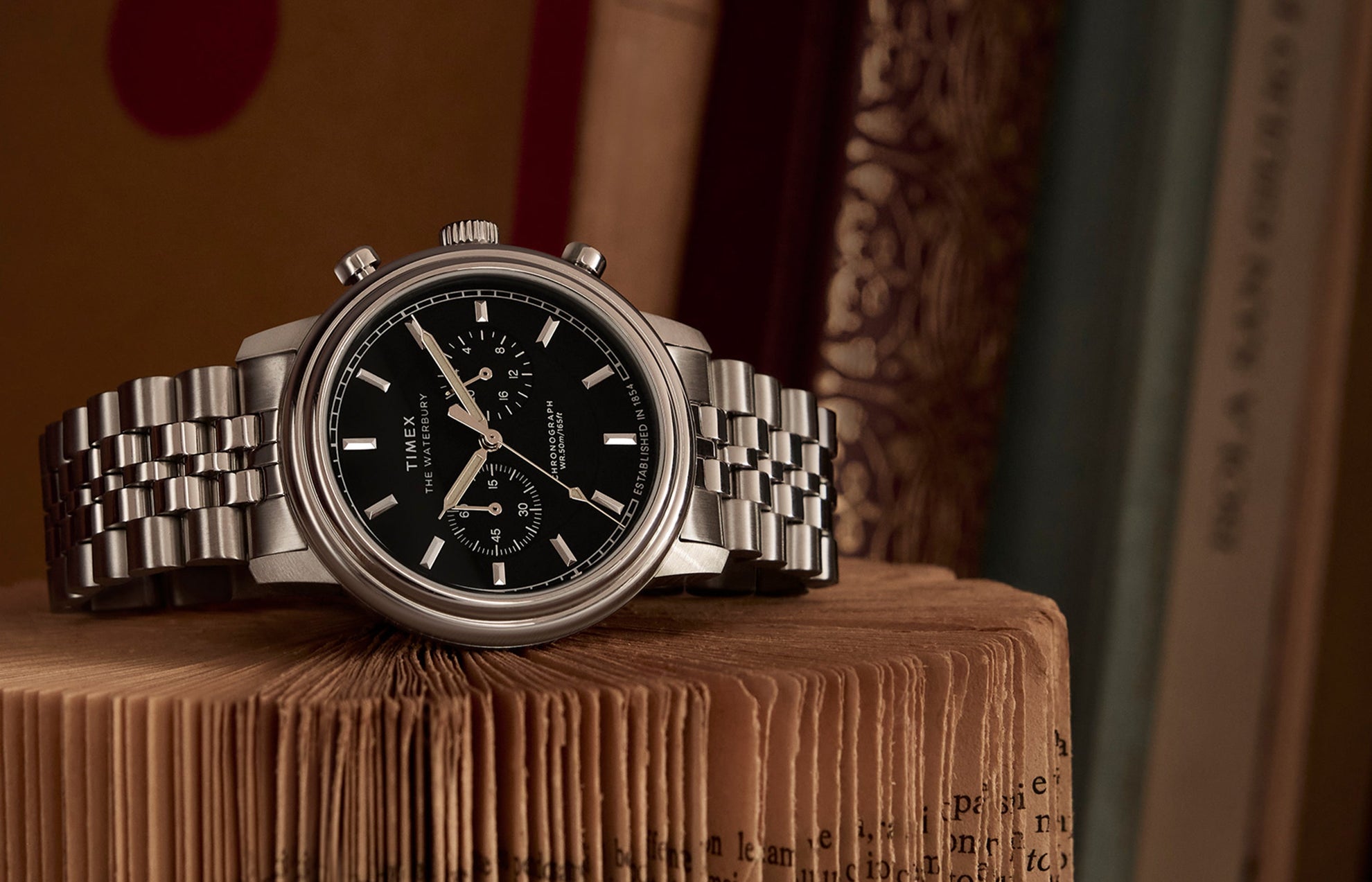 Timex Waterbury Metropolitan Chronograph with black dial and stainless steel bracelet resting on an open vintage book, showcasing its polished finish and sub-dials.
