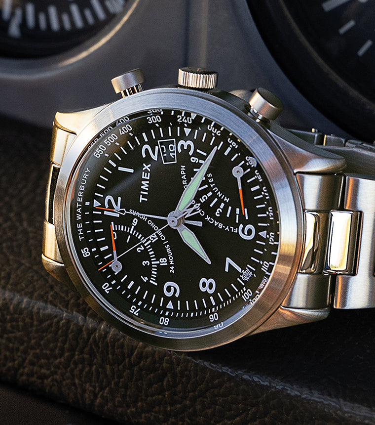 Timex Waterbury Traditional Fly-Back Chronograph resting on an aircraft instrument panel, featuring a black dial, stainless steel bracelet, and luminous hands.