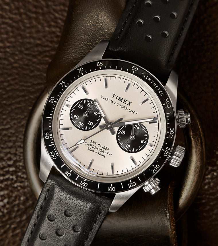 Timex Waterbury Heritage Chronograph with silver dial and black sub-dials resting on brown leather, featuring a black perforated leather strap and tachymeter bezel.