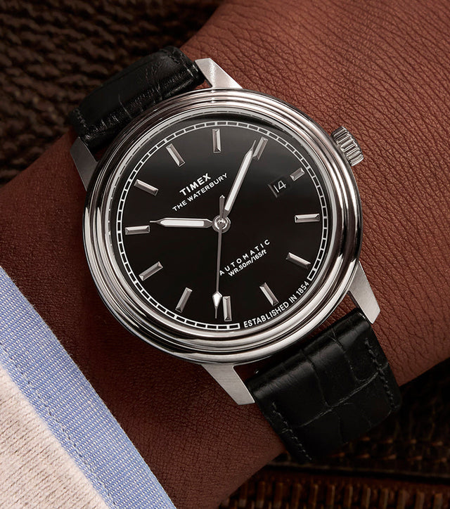Timex Waterbury Metropolitan Automatic watch on wrist, paired with a dress shirt and jacket, showcasing the sleek black dial and classic leather strap.