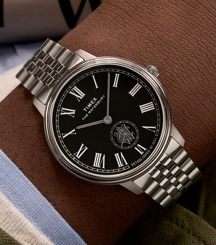 Timex Waterbury Metropolitan Sub-Second watch with black dial, Roman numerals, and stainless steel bracelet worn on wrist with a green jacket.