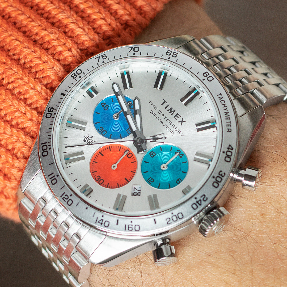 Timex Waterbury Chronograph on wrist with silver-tone stainless steel bracelet, featuring a silver dial and vibrant red and blue sub-dials under natural light.
