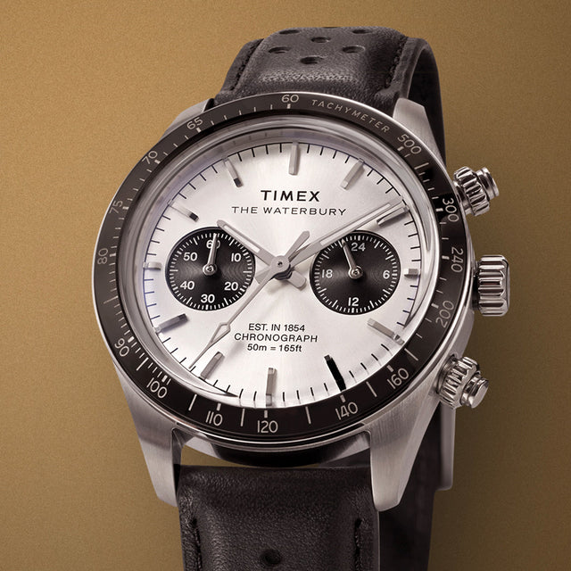 Close-up of the Timex Waterbury Heritage Chronograph showing polished stainless steel case, silver dial, and tachymeter bezel on a gold background.
