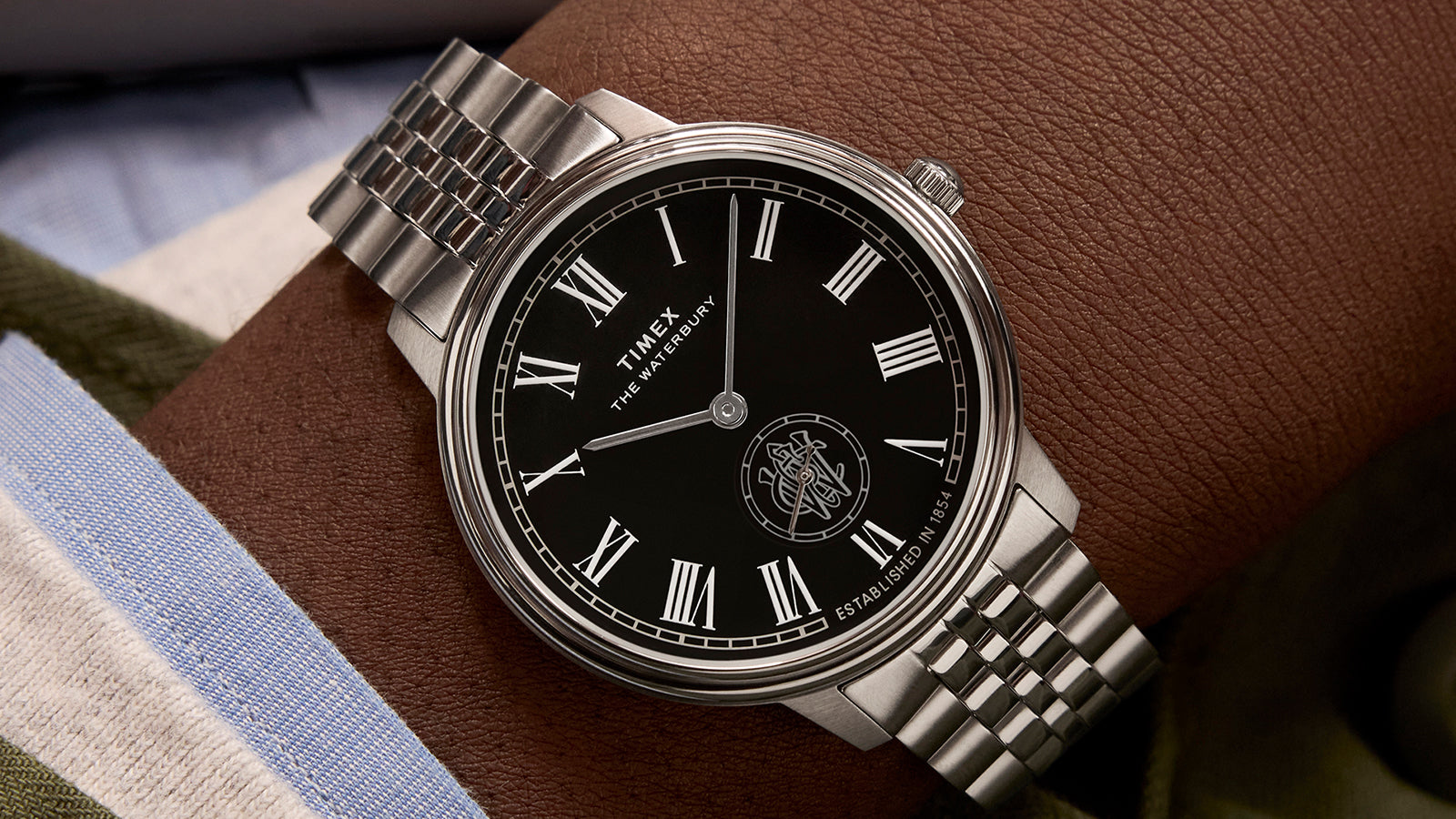 Timex Waterbury Metropolitan Sub-Second watch with black dial, Roman numerals, and stainless steel bracelet worn on wrist with a green jacket.