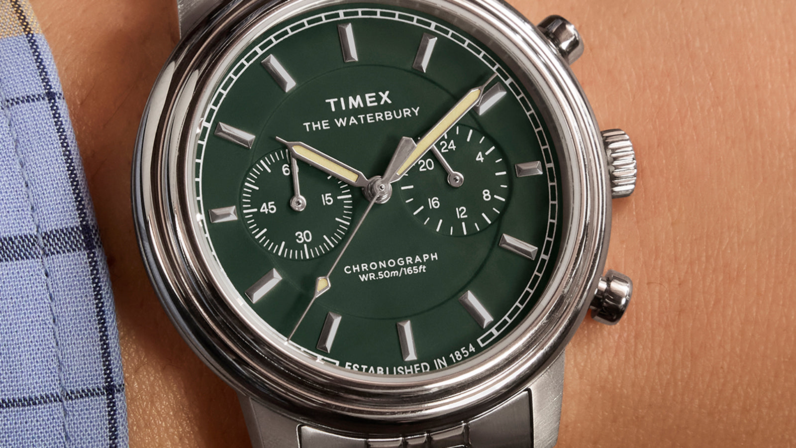 Timex Waterbury Metropolitan Chronograph with green dial and stainless steel bracelet worn on wrist, paired with a plaid shirt and classic styling.