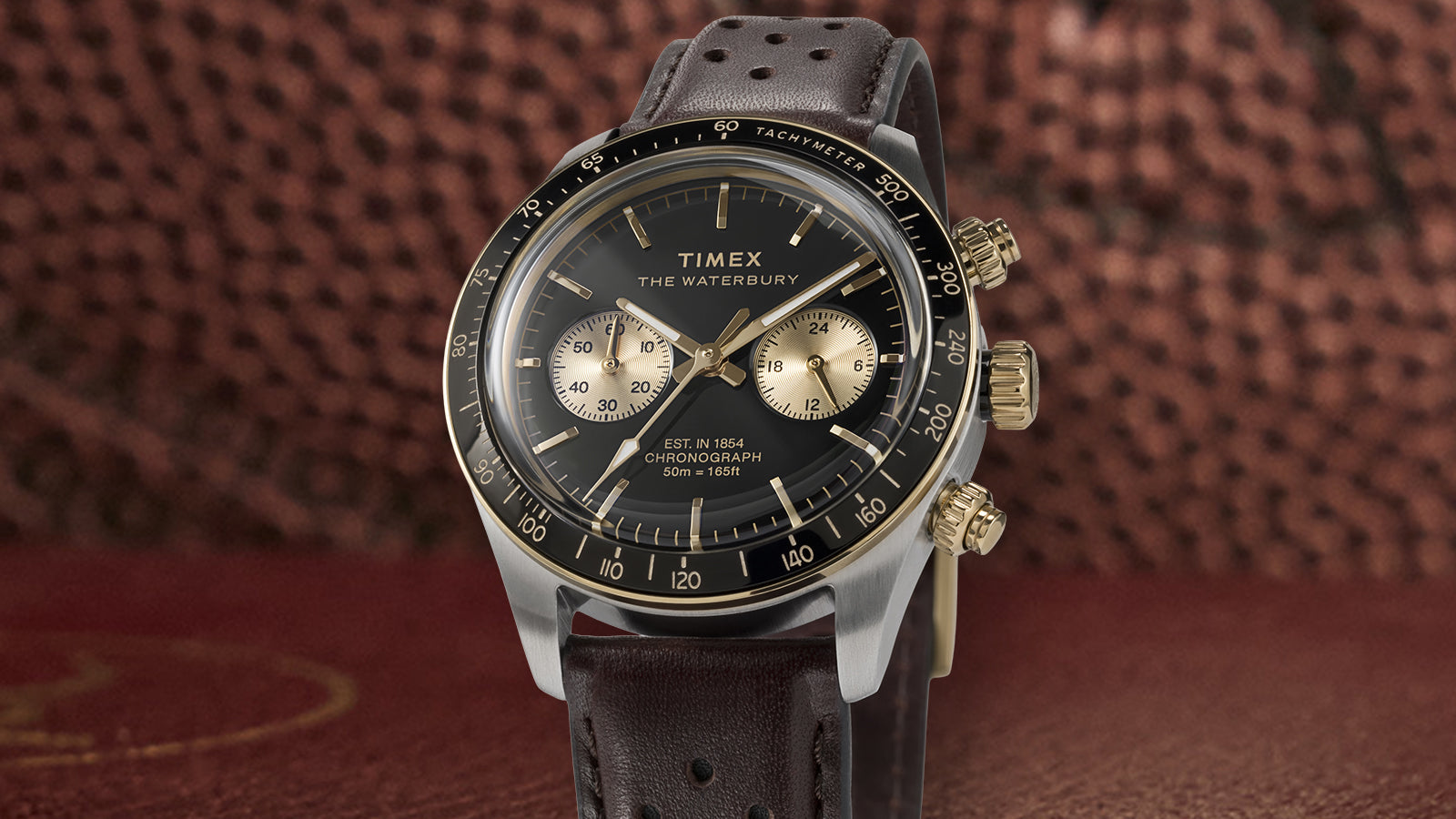 Front-facing view of the Timex Waterbury Heritage Chronograph featuring a black dial, gold-tone accents, and brown perforated leather strap.