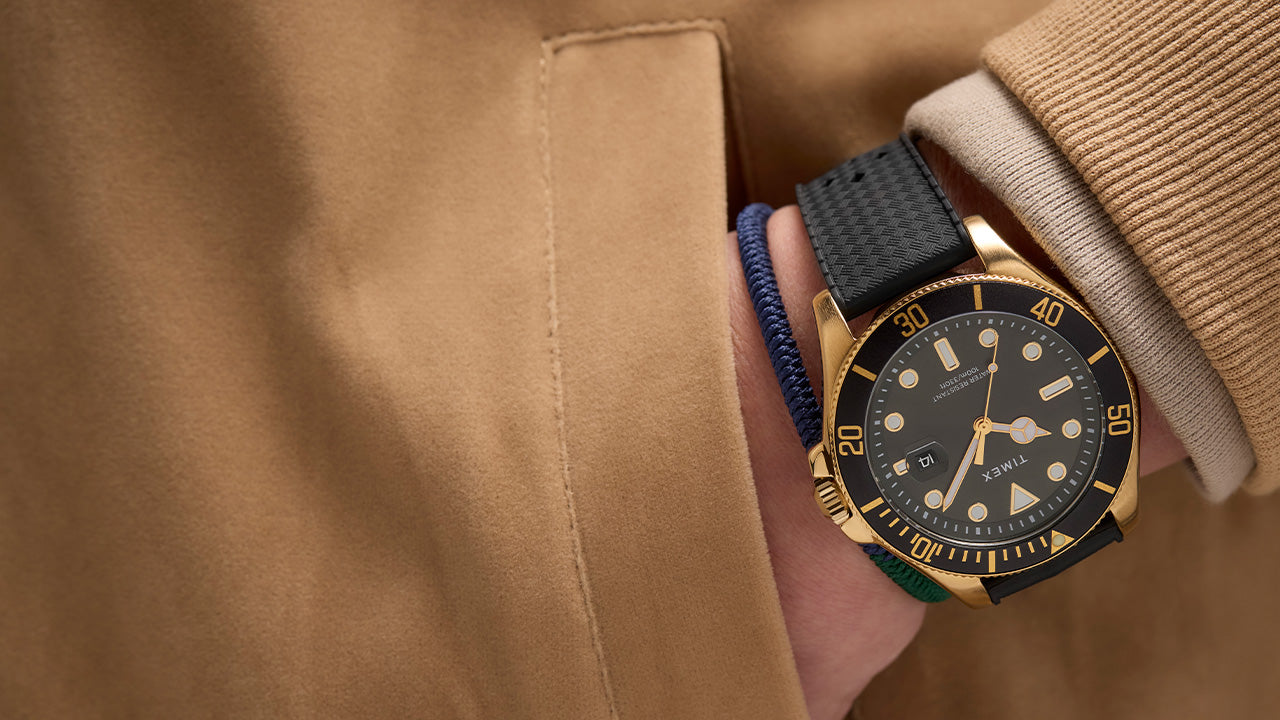 Timex Harborside Coast gold-tone watch with black bezel and textured black strap, styled with a tan jacket and navy braided bracelet on wrist.