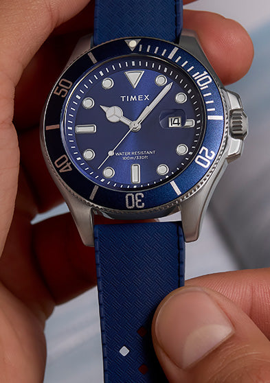 Close-up of a person holding a Timex Harborside Coast watch with a blue dial and bezel, and blue textured strap, near an orange vintage camera.