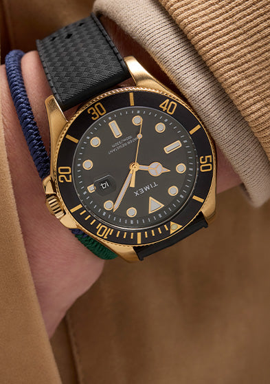 Timex Harborside Coast gold-tone watch with black bezel and textured black strap, styled with a tan jacket and navy braided bracelet on wrist.