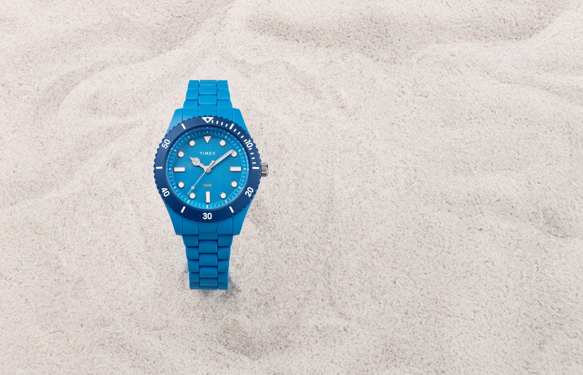 The TIMEX® Legacy Ocean #tide watch in bright blue rests on pale sand, featuring a wave-pattern dial and blue rotating bezel.