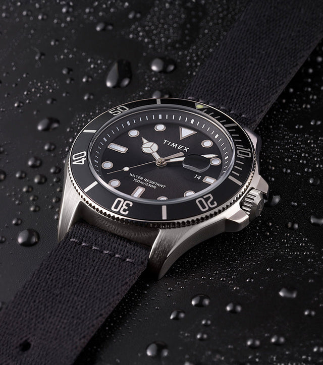 TIMEX® Harborside Coast watch with black dial and black fabric strap resting on a water-dappled black surface, showcasing its water-resistant design.