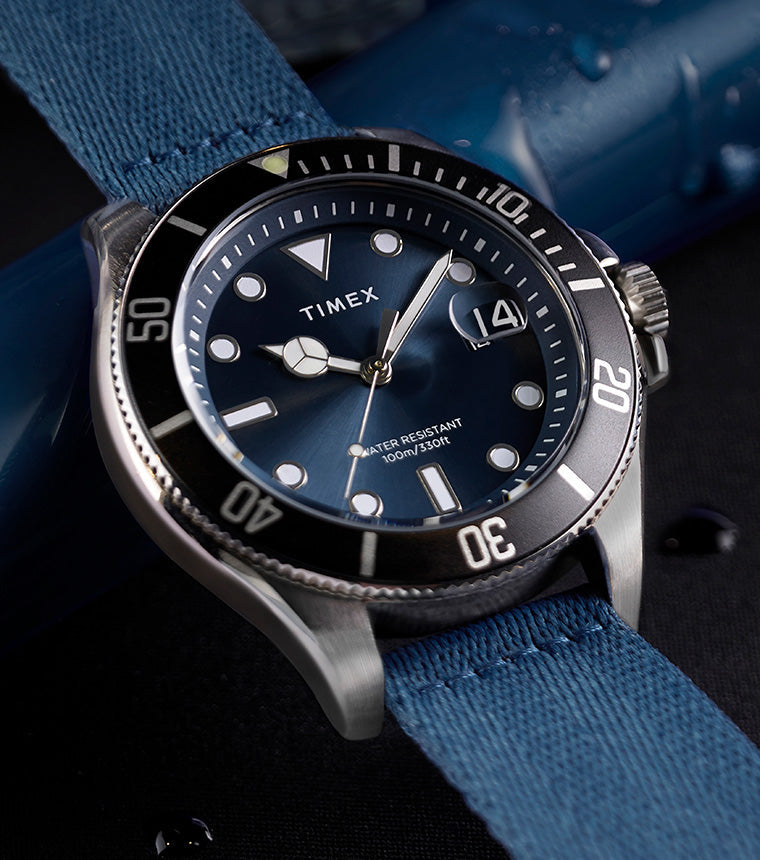 TIMEX® Harborside Coast watch with blue dial, silver-tone case, and blue fabric strap resting on a wet surface with water droplets.