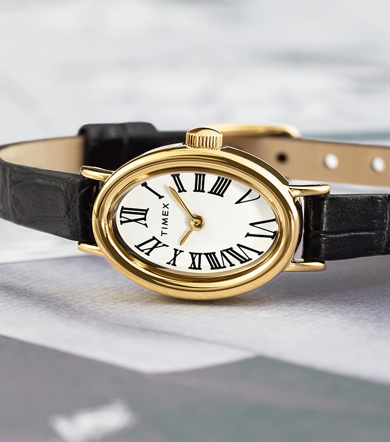 TIMEX® Cavatina watch with gold-tone oval case, white dial, and black leather strap resting on an open magazine.