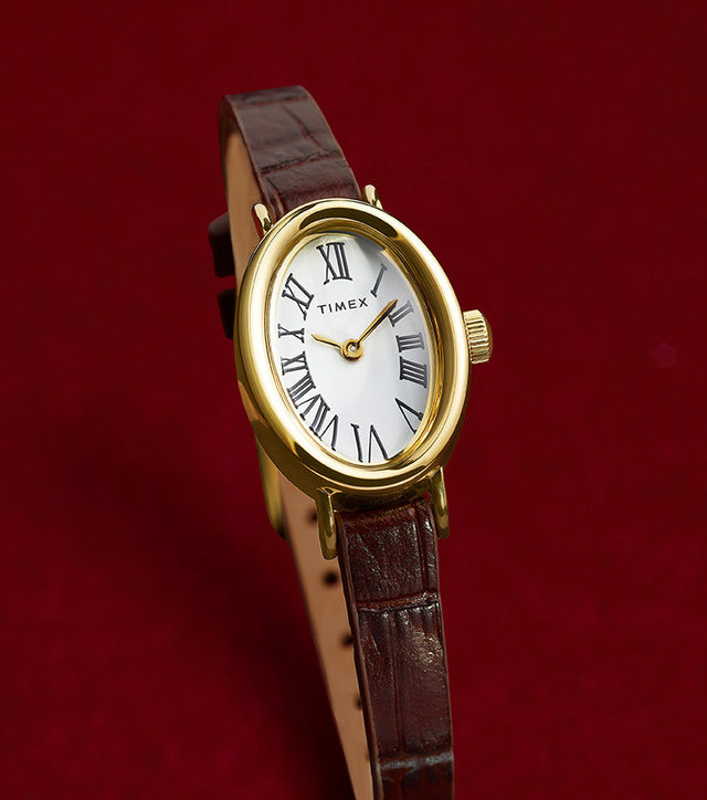 TIMEX® Cavatina watch with gold-tone case and brown leather strap displayed on a rich red background, highlighting its elegant silhouette.